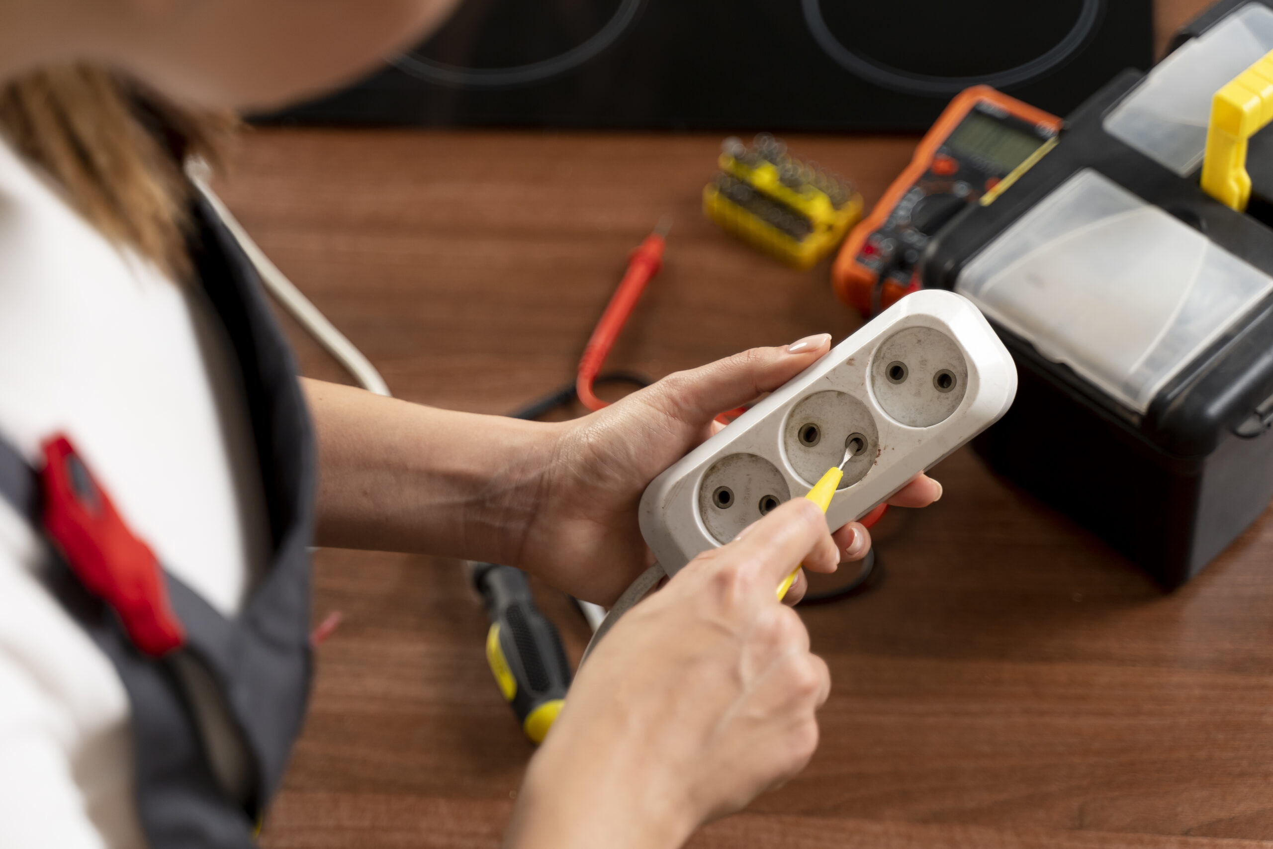 A person uses a screwdriver to work on a multi-outlet power strip on a wooden surface, with electrical tools and a tester visible nearby.